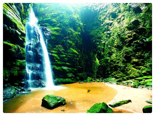 Vista de baixo para cima de uma gruta, com paredes cheias de musgo verde, uma pequena cachoeira e seu chão todo formado de arreia, com algumas rochas.